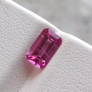 Puede incluir: Una gema rosa vibrante con un corte rectangular. La gema está colocada sobre un fondo blanco.