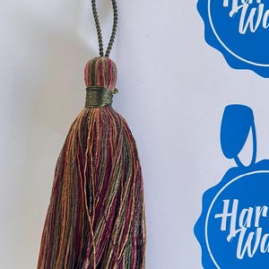 May include: A decorative tassel with a loop for hanging. The tassel features a mix of colors including burgundy, gold, green, and brown. The top of the tassel is wrapped in green thread.