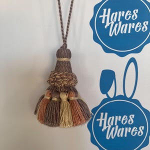 May include: A decorative tassel with a braided cord in shades of brown, tan, and peach. The tassel features a woven top section and multiple strands of yarn, perfect for home decor or craft projects. The image also shows the text "Hares Wares".