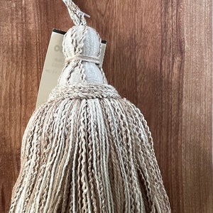 Puede incluir: Un borla beige y marrón con un cordón largo y una llave antigua adjunta. La borla está hecha de un material suave y esponjoso y tiene un nudo decorativo en la parte superior.