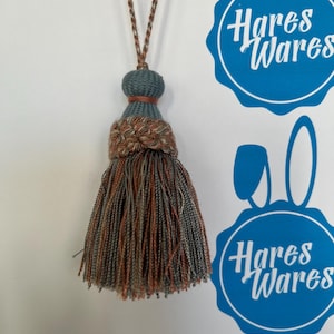 Könnte beinhalten: Ein dekorativer Quastenanhänger mit einem gedrehten Kordel in Braun-, Grau- und Orangetönen. Die Quaste hat einen teal-farbenen Oberteil und einen gewebten Abschnitt in Braun und Orange. Das Bild enthält auch ein blaues Logo mit der Aufschrift "Hares Wares".