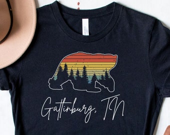 May include: Black t-shirt with a vintage-style graphic of a bear silhouette with a sunset behind it and pine trees in front of the bear. The text "Gatlinburg, TN" is printed in white below the graphic.