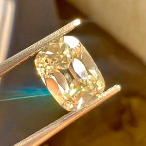 K&ouml;nnte beinhalten: Ein hellgelber Diamant im Kissenschliff wird von einer Pinzette gehalten. Der Diamant hat eine rechteckige Form mit abgerundeten Ecken und Facetten, die das Licht reflektieren. Der Edelstein ist hellgelb.