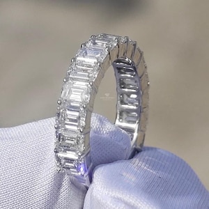 Könnte beinhalten: Ein silberner Ring mit einem durchgehenden Band aus rechteckig geschliffenen, klaren Edelsteinen. Der Ring ist in einem polierten Silberband gefasst, wobei die Edelsteine gleichmäßig über den gesamten Umfang verteilt sind. Der Ring wird vor einem weichen, hellen Stoff präsentiert.