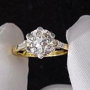 May include: A gold ring with a large, clear, round-cut diamond as the centerpiece. The ring has smaller diamonds on the band and is held by a gloved hand against a black background.