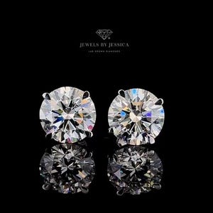 May include: A pair of round-cut stud earrings featuring clear, faceted stones that reflect light with a brilliant sparkle. Above the earrings, the text JEWELS BY JESSICA LAB GROWN DIAMONDS is printed in white against a solid black background.