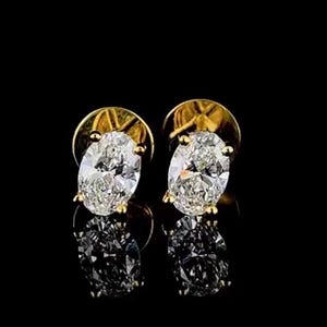 May include: A pair of gold stud earrings featuring oval-cut diamonds. The diamonds are clear and sparkling, set in a gold bezel. The earrings are shown against a black background, with their reflections visible.