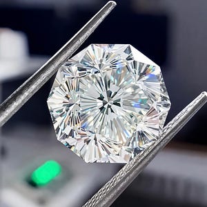 May include: A faceted, brilliant-cut diamond held by a pair of silver tweezers. The gemstone has a complex, symmetrical pattern of facets, reflecting light. The background is blurred, with a hint of green light.