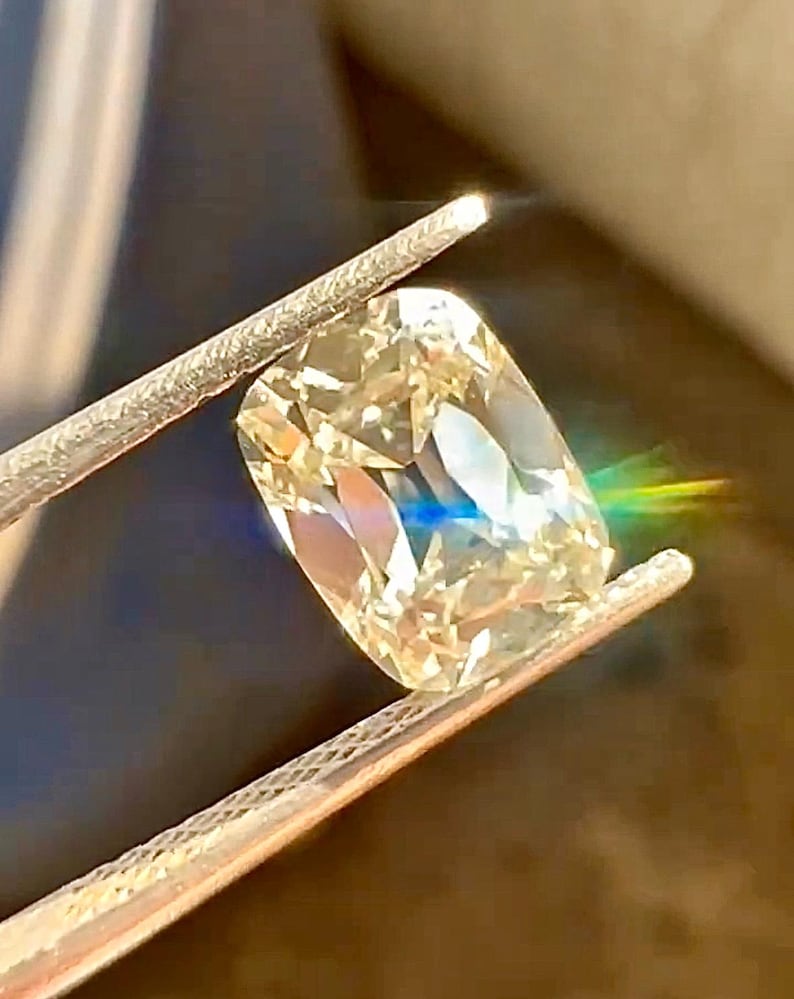 K&ouml;nnte beinhalten: Ein kissenf&ouml;rmiger Diamant, der von einer Pinzette gehalten wird. Der Diamant hat einen warmen, gelblichen Farbton und reflektiert das Licht, wodurch ein Regenbogeneffekt entsteht. Die Pinzette ist silberfarben, und der Hintergrund ist unscharf.