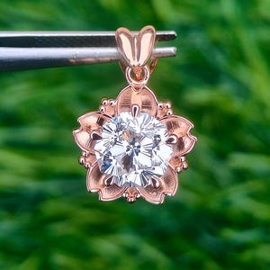 May include: Rose gold pendant featuring a sparkling, sakura-cut gemstone set within a flower-shaped frame. The pendant has a delicate heart-shaped bail. The background is a blurred green.