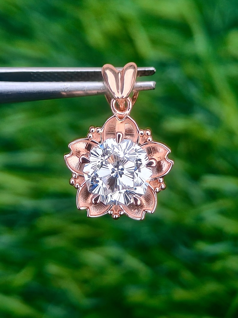 May include: Rose gold pendant featuring a sparkling, sakura-cut gemstone set within a flower-shaped frame. The pendant has a delicate heart-shaped bail. The background is a blurred green.