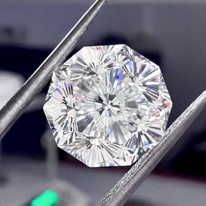 May include: A brilliant-cut, octagonal diamond held by silver tweezers. The gemstone has many facets, reflecting light and creating a sparkling effect. The diamond is clear and appears flawless, showcasing its intricate design and craftsmanship.