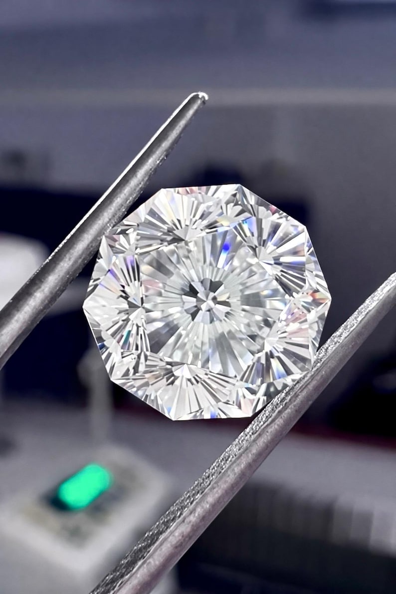 May include: A brilliant-cut, octagonal diamond held by silver tweezers. The gemstone has many facets, reflecting light and creating a sparkling effect. The diamond is clear and appears flawless, showcasing its intricate design and craftsmanship.