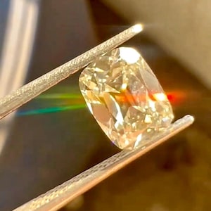 K&ouml;nnte beinhalten: Ein gro&szlig;er Diamant im Kissenschliff, der von einer Pinzette gehalten wird. Der Diamant hat einen warmen, gelben Farbton und reflektiert das Licht, wodurch ein Regenbogeneffekt entsteht. Die Pinzette ist silberfarben, und der Hintergrund ist unscharf.
