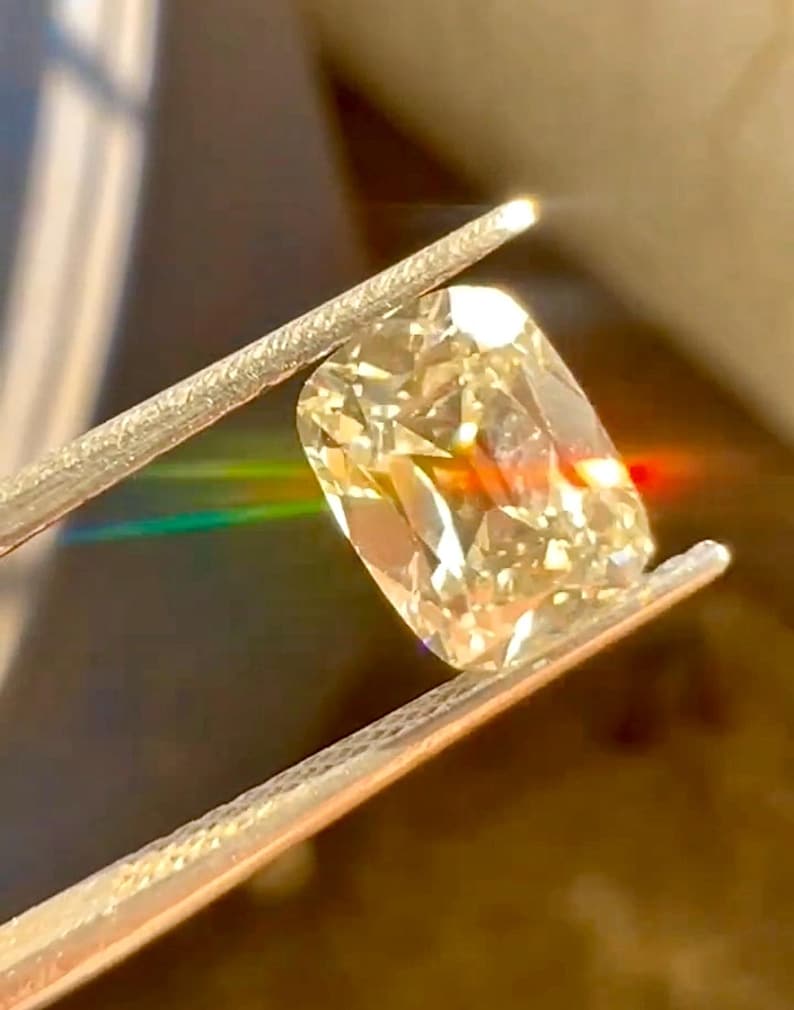 K&ouml;nnte beinhalten: Ein gro&szlig;er Diamant im Kissenschliff, der von einer Pinzette gehalten wird. Der Diamant hat einen warmen, gelben Farbton und reflektiert das Licht, wodurch ein Regenbogeneffekt entsteht. Die Pinzette ist silberfarben, und der Hintergrund ist unscharf.