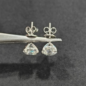 May include: A pair of silver-toned stud earrings featuring clear, faceted, round-cut stones held in four-prong settings. The earrings include matching silver-toned butterfly-style earring backs, shown held together by a pair of metal tweezers.