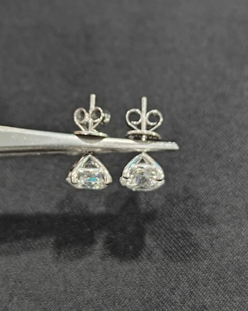 May include: A pair of silver-toned stud earrings featuring clear, faceted, round-cut stones held in four-prong settings. The earrings include matching silver-toned butterfly-style earring backs, shown held together by a pair of metal tweezers.