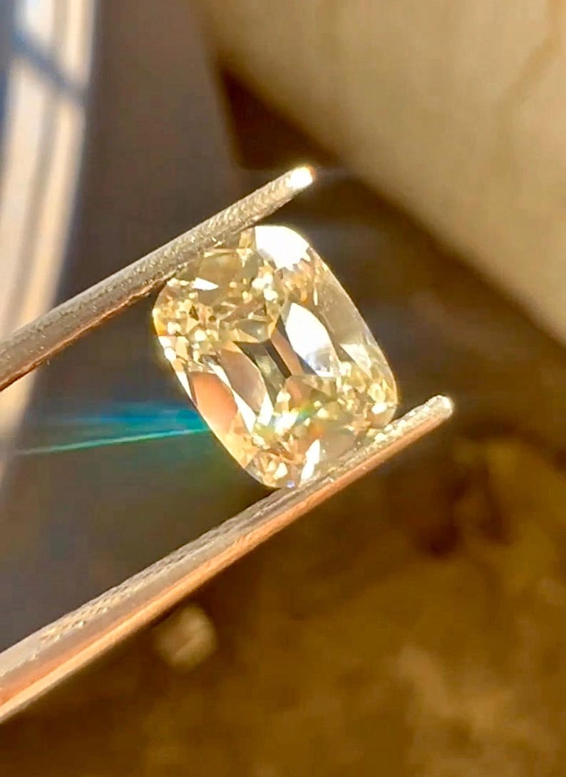 K&ouml;nnte beinhalten: Ein hellgelber Diamant im Kissenschliff wird von einer Pinzette gehalten. Der Diamant hat eine rechteckige Form mit abgerundeten Ecken und Facetten, die das Licht reflektieren. Der Edelstein ist hellgelb.