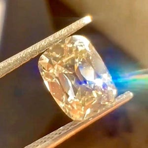 K&ouml;nnte beinhalten: Nahaufnahme eines kissenf&ouml;rmigen Diamanten, der von einer Pinzette gehalten wird. Der Diamant hat eine warme Champagnerfarbe und reflektiert das Licht, wodurch ein brillantes Funkeln entsteht. Das Bild zeigt die Facetten und die Klarheit des Edelsteins.