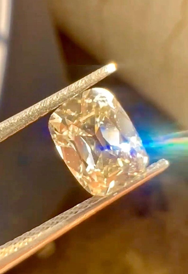 K&ouml;nnte beinhalten: Nahaufnahme eines kissenf&ouml;rmigen Diamanten, der von einer Pinzette gehalten wird. Der Diamant hat eine warme Champagnerfarbe und reflektiert das Licht, wodurch ein brillantes Funkeln entsteht. Das Bild zeigt die Facetten und die Klarheit des Edelsteins.