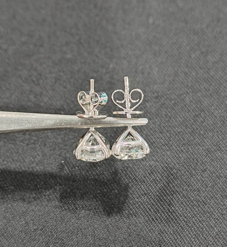 May include: A pair of silver-toned stud earrings featuring round, faceted clear stones set in a four-prong basket setting. The earrings include butterfly-style friction backs with a heart-shaped wire design, held together by metal tweezers.