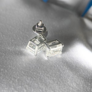 K&ouml;nnte beinhalten: Ein Paar silberne Ohrringe mit einem quadratischen Diamanten in jedem Ohrring.