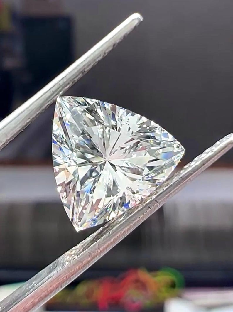 Pu&ograve; includere: Un diamante a taglio triangolare, tenuto da pinzette d'argento. La gemma brilla con numerose sfaccettature, riflettendo la luce. L'immagine &egrave; un primo piano che evidenzia la chiarezza e il taglio del diamante.