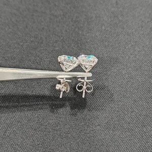 May include: A pair of silver-toned stud earrings featuring round, faceted clear stones set in four-prong baskets. The earrings are held by metal tweezers, displaying the detailed metal posts and the intricate, swirling wire butterfly-style earring backs.