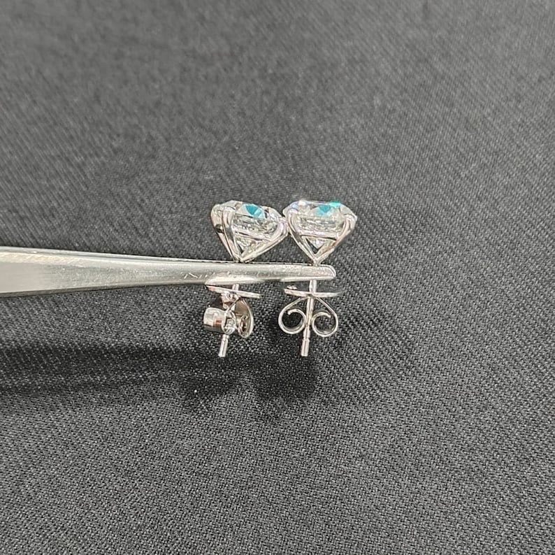 May include: A pair of silver-toned stud earrings featuring round, faceted clear stones set in four-prong baskets. The earrings are held by metal tweezers, displaying the detailed metal posts and the intricate, swirling wire butterfly-style earring backs.