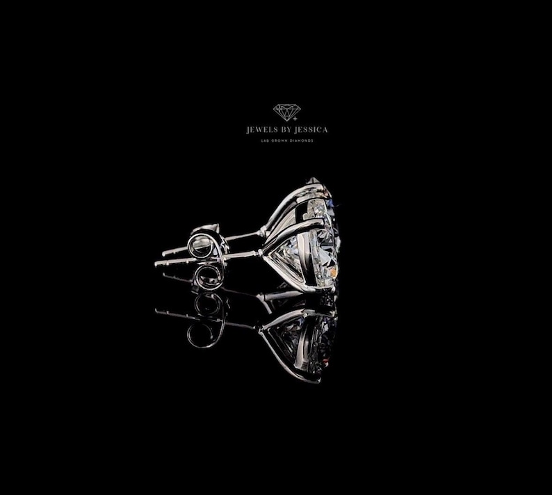 May include: A silver-toned stud earring featuring a large, clear, faceted gemstone set in a multi-prong basket setting. The text JEWELS BY JESSICA LAB GROWN DIAMONDS is visible above the earring, which is reflected on the black surface.