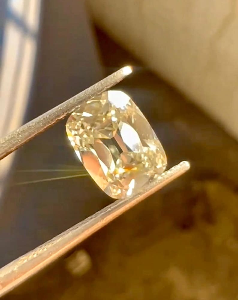 K&ouml;nnte beinhalten: Ein hellgelber Diamant im Kissenschliff wird von einer Pinzette gehalten. Der Diamant hat viele Facetten, die das Licht reflektieren und einen funkelnden Effekt erzeugen. Der Hintergrund ist verschwommen und warm get&ouml;nt.