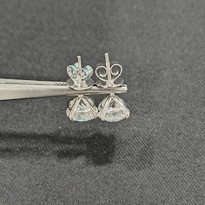 May include: A pair of silver-toned stud earrings featuring clear, round-cut faceted stones held in four-prong settings. The earrings include butterfly-style friction backs and are displayed side-by-side, showcasing the metal prongs and the clear, reflective quality of the stones.