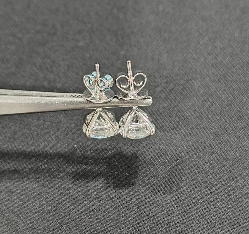 May include: A pair of silver-toned stud earrings featuring clear, round-cut faceted stones held in four-prong settings. The earrings include butterfly-style friction backs and are displayed side-by-side, showcasing the metal prongs and the clear, reflective quality of the stones.