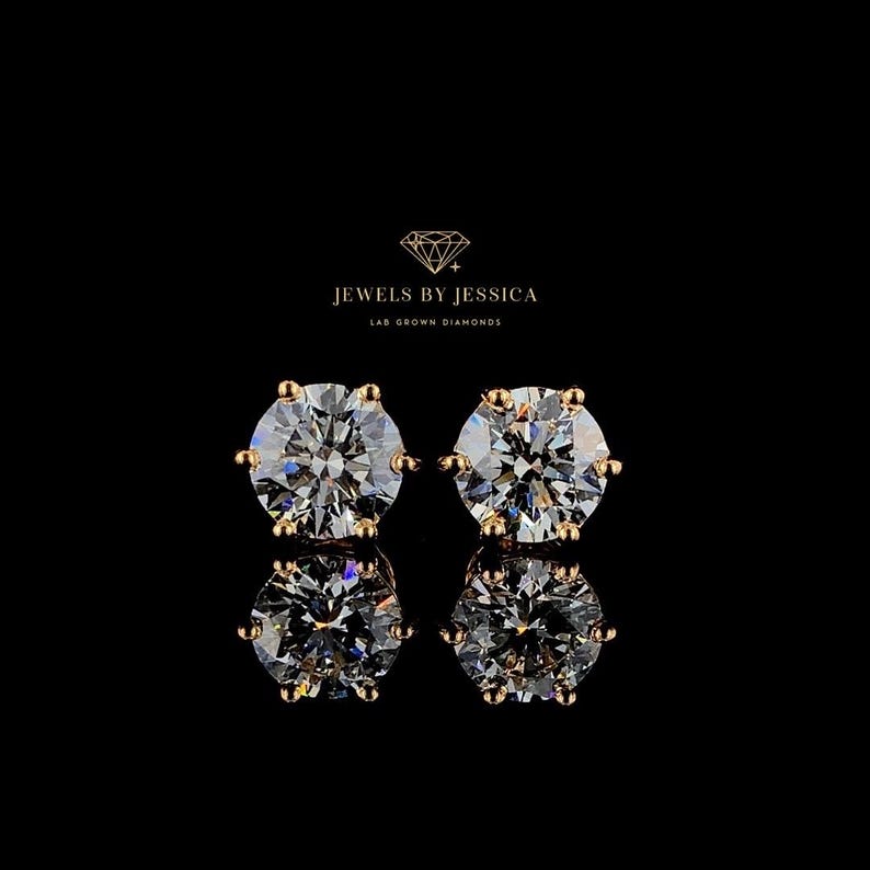 May include: A pair of gold stud earrings with round, clear lab-grown diamonds. The earrings are set in a classic prong setting. "Jewels by Jessica Lab Grown Diamonds" is written in the top center of the image.