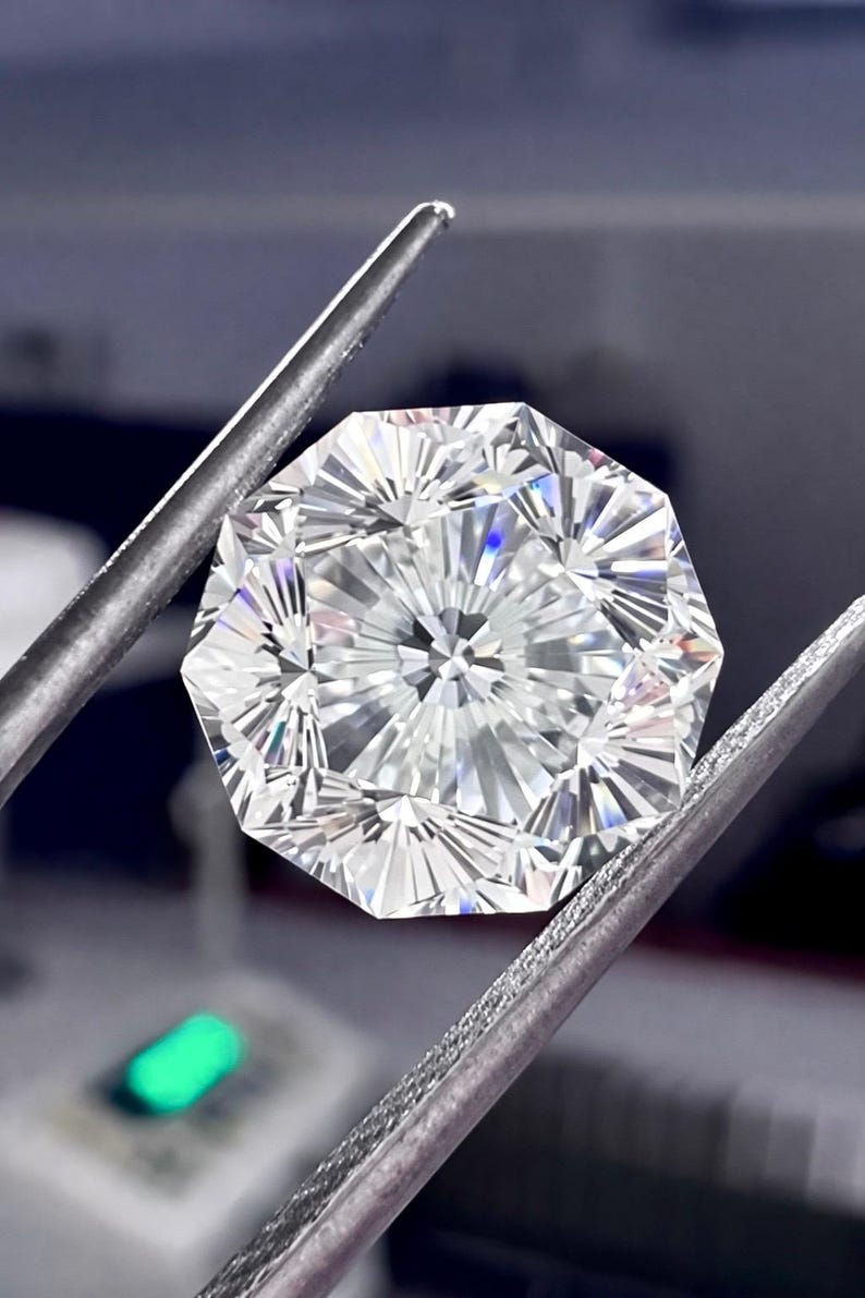 May include: A brilliant-cut, octagonal diamond held by silver tweezers. The gemstone has many facets, reflecting light and creating a sparkling effect. The diamond is clear and appears flawless, showcasing its intricate design and craftsmanship.