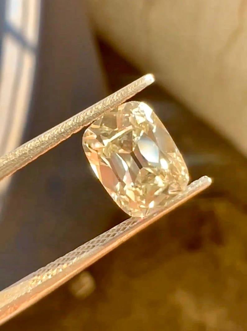 K&ouml;nnte beinhalten: Ein kissenf&ouml;rmiger, hellgelber Diamant, der von einer Pinzette gehalten wird. Der Edelstein ist rechteckig mit abgerundeten Ecken und reflektiert das Licht, wodurch seine Facetten zur Geltung kommen. Die Pinzette ist silberfarben, und der Hintergrund ist verschwommen und warm get&ouml;nt.