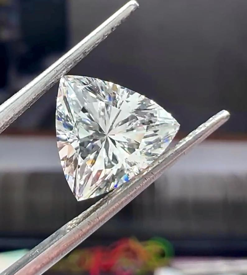 Pu&ograve; includere: Un diamante a taglio brillante, di forma triangolare, tenuto da pinzette d'argento. La gemma ha molte sfaccettature, che riflettono la luce e creano un effetto scintillante. Il diamante ha una forma triangolare.