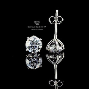 May include: A pair of silver-toned stud earrings featuring round, faceted clear stones set in a four-prong basket setting. The text JEWELS BY JESSICA LAB GROWN DIAMONDS is visible above the earrings. The earrings are shown from the front and side.