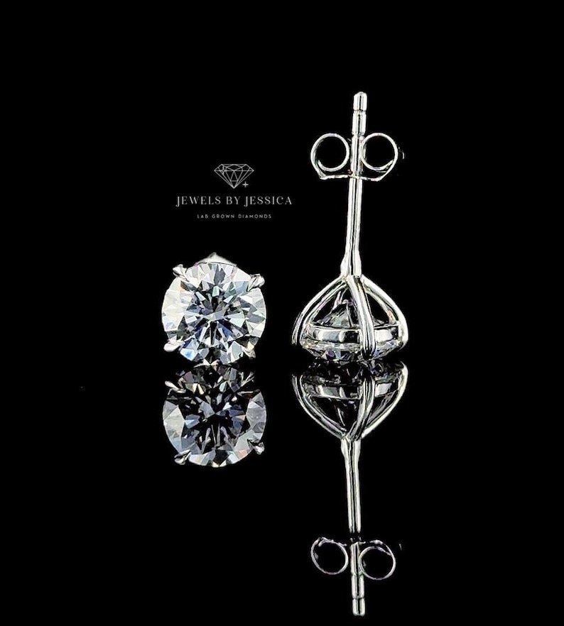May include: A pair of silver-toned stud earrings featuring round, faceted clear stones set in a four-prong basket setting. The text JEWELS BY JESSICA LAB GROWN DIAMONDS is visible above the earrings. The earrings are shown from the front and side.