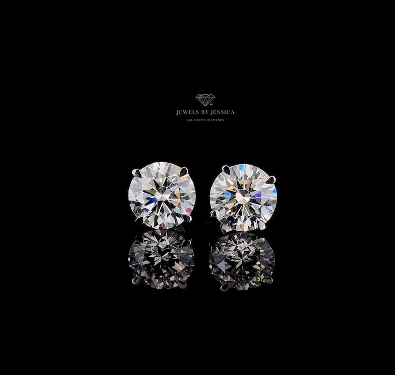 May include: A pair of round-cut stud earrings featuring clear, faceted stones that reflect light with a brilliant sparkle. Above the earrings, the text JEWELS BY JESSICA LAB GROWN DIAMONDS is printed in white against a solid black background.