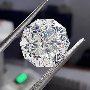 May include: A faceted, octagonal-cut diamond held by a pair of silver tweezers. The diamond is clear and reflects light, showcasing its many facets. The background is blurred, with a hint of green light.