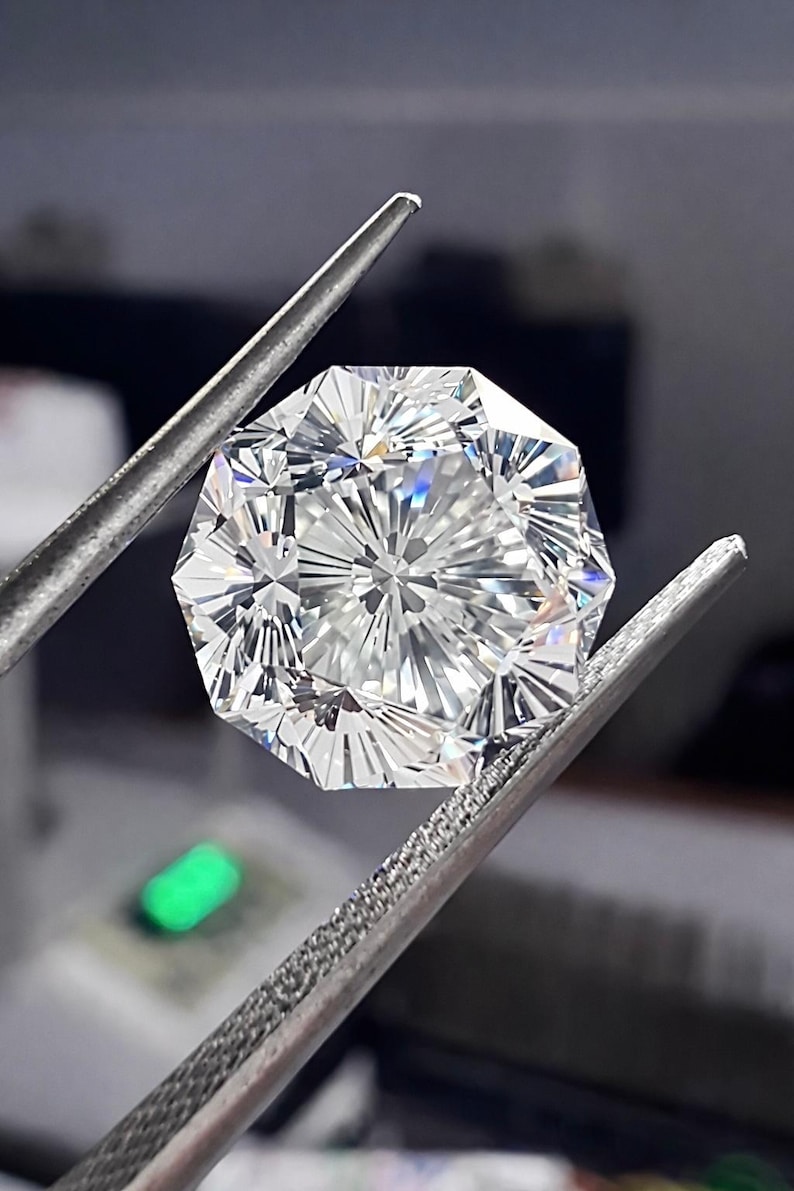 May include: A faceted, octagonal-cut diamond held by a pair of silver tweezers. The diamond is clear and reflects light, showcasing its many facets. The background is blurred, with a hint of green light.
