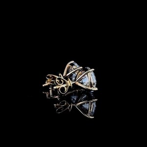 May include: A pair of stud earrings featuring round, clear, faceted gemstones set in gold-toned metal prongs. The earrings are shown from a side angle, highlighting the basket setting and the metal post backs against a solid black background.