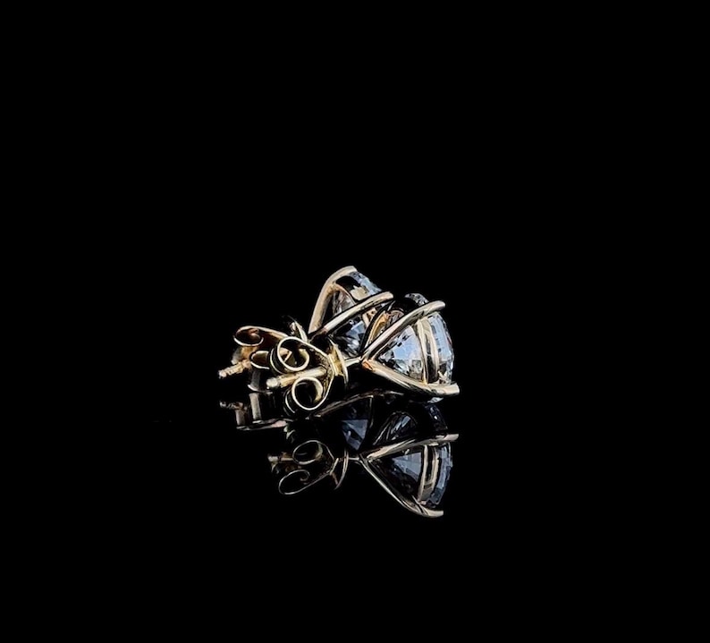 May include: A pair of stud earrings featuring round, clear, faceted gemstones set in gold-toned metal prongs. The earrings are shown from a side angle, highlighting the basket setting and the metal post backs against a solid black background.