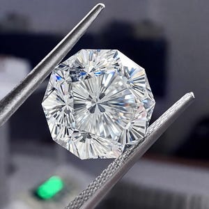 May include: A faceted, clear gemstone with an octagonal shape is held by a pair of silver tweezers. The gem has intricate internal facets that reflect light, creating a brilliant display. The background is blurred.