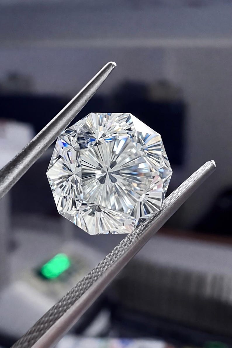 May include: A faceted, clear gemstone with an octagonal shape is held by a pair of silver tweezers. The gem has intricate internal facets that reflect light, creating a brilliant display. The background is blurred.