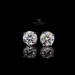 May include: A pair of round brilliant cut stud earrings with clear, faceted stones set in four-prong metal settings. The text JEWELS BY JESSICA LAB GROWN DIAMONDS is visible above the earrings, which are reflected on a black surface.