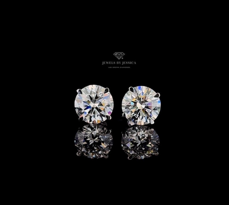 May include: A pair of round brilliant cut stud earrings with clear, faceted stones set in four-prong metal settings. The text JEWELS BY JESSICA LAB GROWN DIAMONDS is visible above the earrings, which are reflected on a black surface.