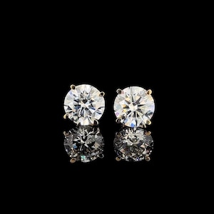 May include: A pair of round-cut stud earrings featuring clear, faceted stones set in gold-toned metal prongs. Each earring displays a brilliant, reflective surface with multiple facets that catch the light, creating a sparkling appearance against a black background.