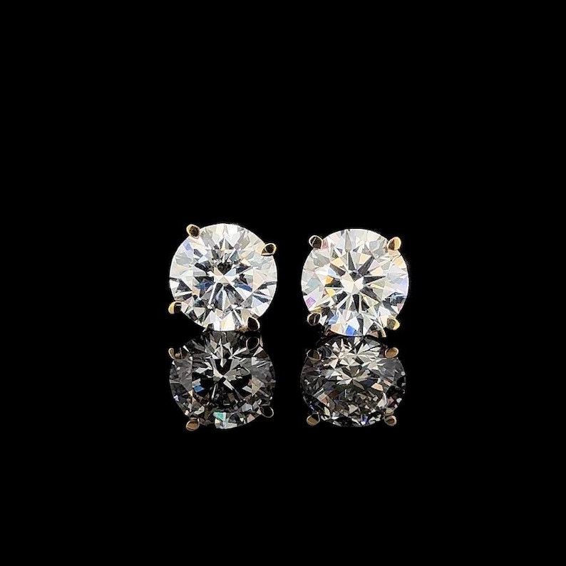 May include: A pair of round-cut stud earrings featuring clear, faceted stones set in gold-toned metal prongs. Each earring displays a brilliant, reflective surface with multiple facets that catch the light, creating a sparkling appearance against a black background.